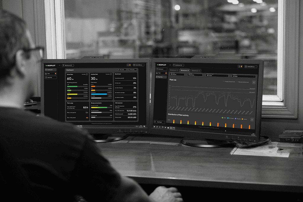 Manager in office overlooking factory and computer screens showing UgoPilot fleet management software dashboard and reporting