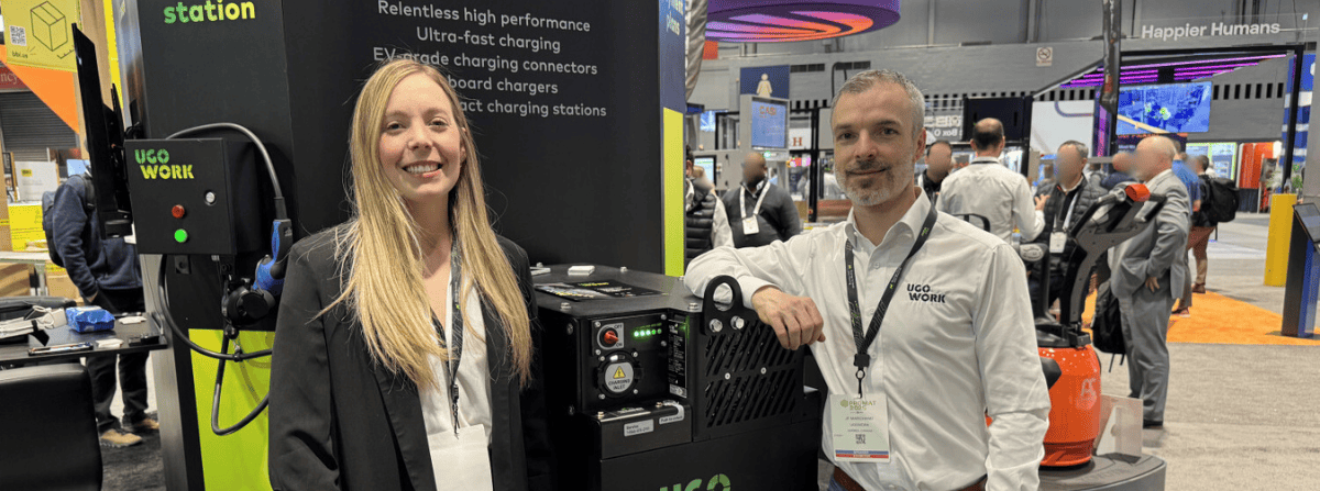 Two smiling UgoWork team members standing next to a UgoWork lithium-ion battery on display at the ProMat trade show.