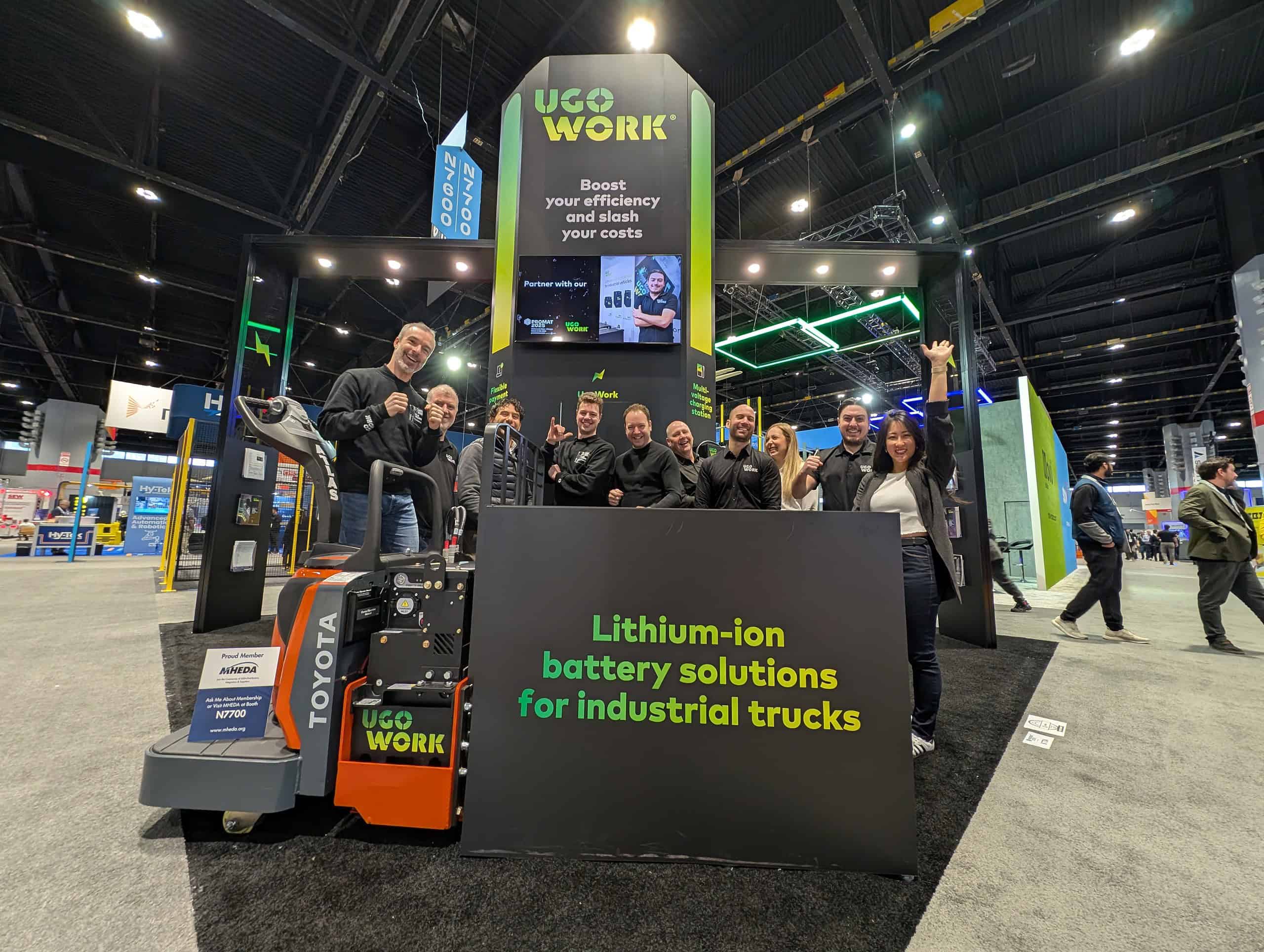 Group photo of the UgoWork team celebrating in front of their booth at PROMAT 2025 with a lithium-ion battery-powered pallet jack.