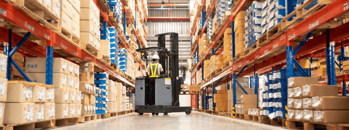 Cariste à l’œuvre dans une allée étroite d’entrepôt, entre des rayonnages chargés de boîtes et de contenants.