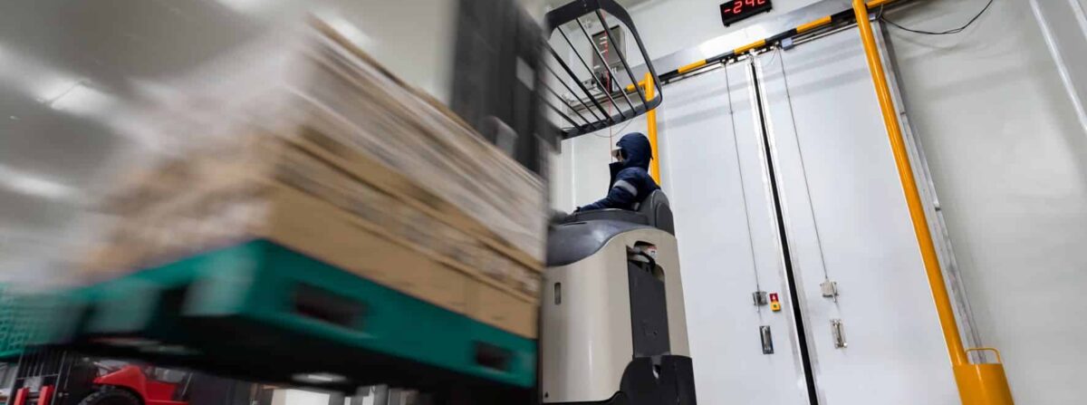 Warehouse worker Stand-on stacker battery-powered truck used to lift and move goods stock in cold room or freezer room.