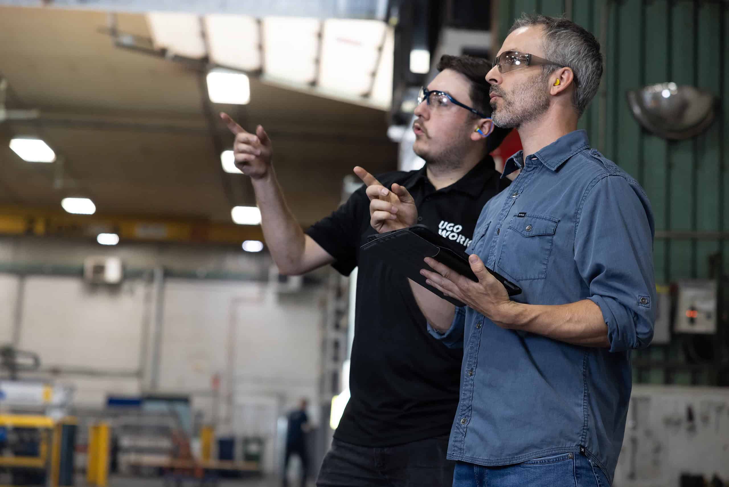 Deux hommes debout dans une usine regardant une tablette et pointant vers l’avant.
