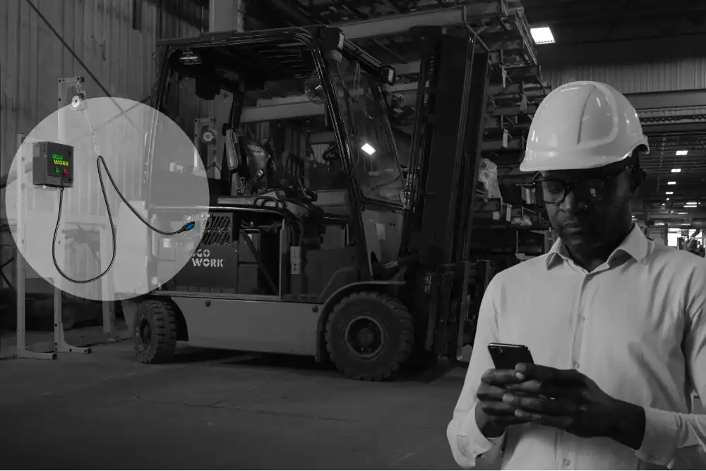Station de recharge compacte UgoWork pour chariot élévateur dans un entrepôt industriel avec un travailleur utilisant son smartphone.