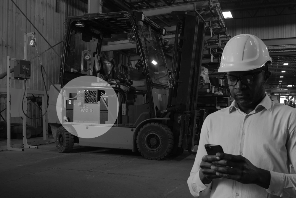 Gestionnaire de flotte utilisant un appareil mobile dans un entrepôt industriel avec, en arrière-plan, un chariot élévateur alimenté par une batterie au lithium UgoWork.