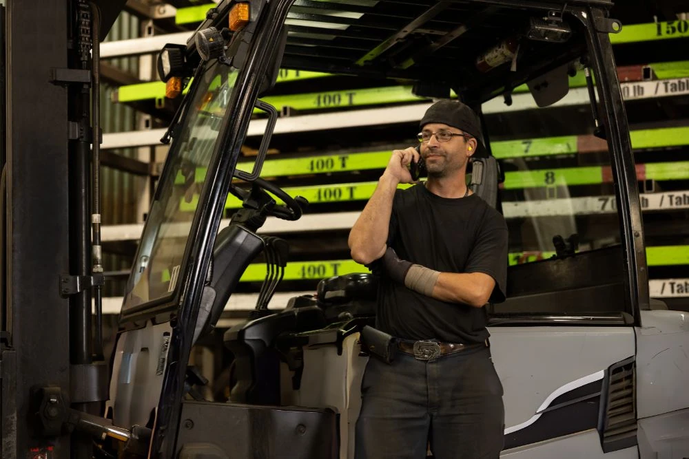 Opérateur de chariot élévateur parlant au téléphone à côté d'un chariot élévateur en panne dans un entrepôt avec des rayonnages industriels.
